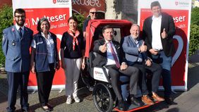 Zwei Männer sitzen in einer Fahrrad-Rikscha und mehrere Menschen stehen darum herum.