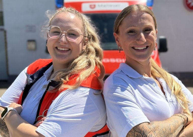 Zwei strahlend lächelnde Rettungsdienst-Mitarbeiterinnen vor einer Rettungswache
