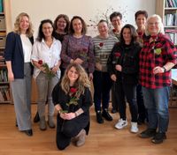 Eine Gruppe von Personen steht in einem Raum vor einer Bücherwand zusammen und hält jeweils eine einzelne rote Rose in der Hand. Eine Person kniet in der ersten Reihe, die übrigen stehen dahinter in zwei Reihen. Der Raum hat einen Holzfußboden, Regale mit Büchern und Ordnern sowie einen schlichten, hellen Hintergrund.