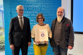 Drei Personen stehen vor einer Wand, eine davon hält eine Urkunde in den Händen.