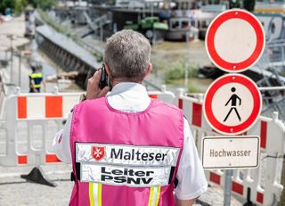 Ein Mann mit einer Weste mit Malteser-Logo telefoniert vor einer abgesperrten Straße