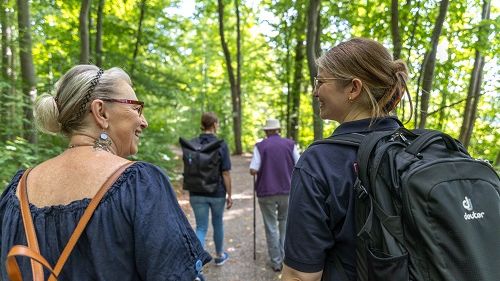 Menschen spazieren durch einen Wald, sie sind von hinten zu sehen. Vorne im Bild sind zwei Frauen, die sich anschauen und sich unterhalten