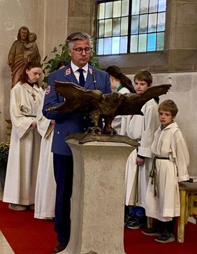 Ein Mann in blauem Uniformanzug steht in einer Kirche neben einem Adler-Standbild, umgeben von Kindern in weißen Gewändern.