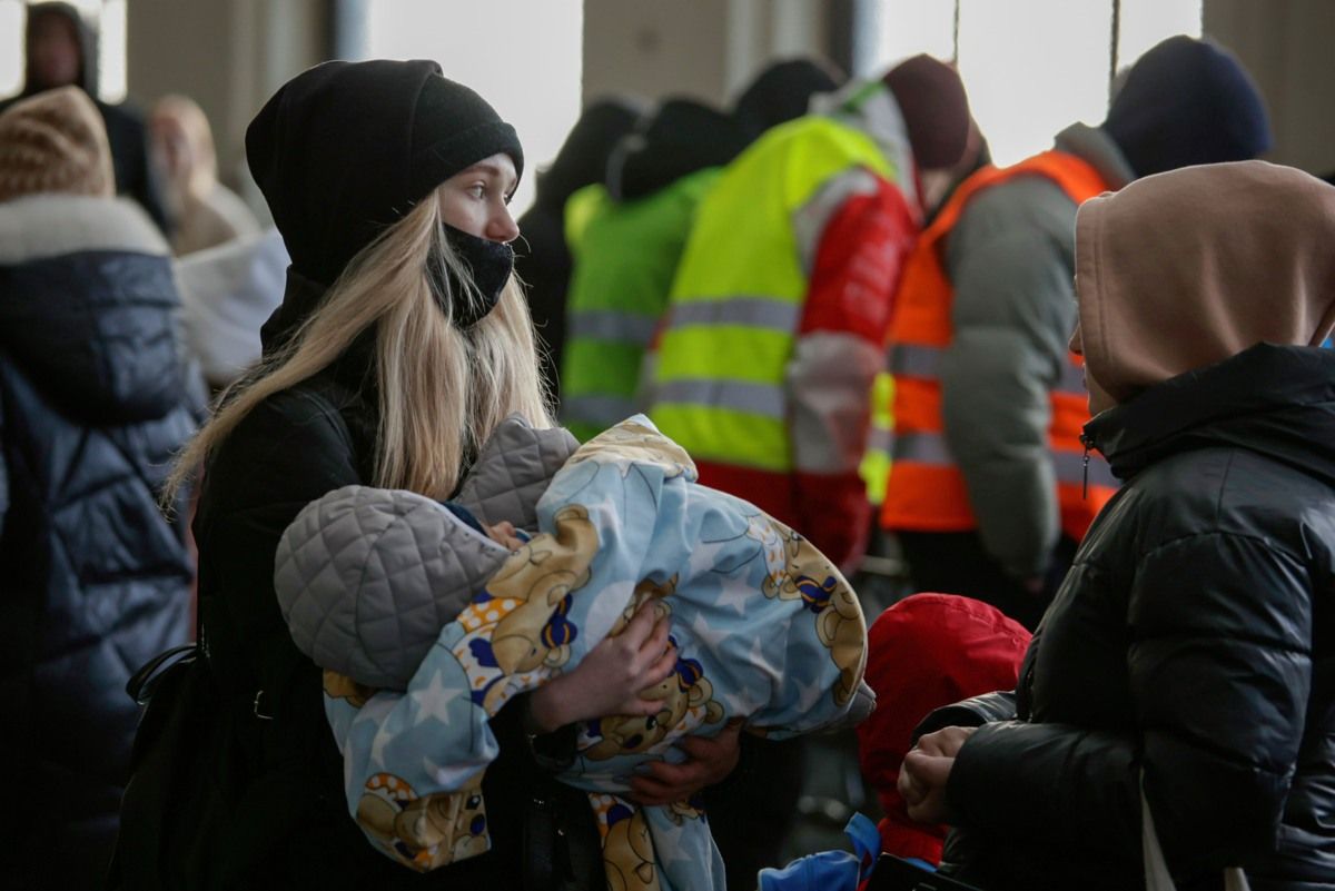 Schwarz gekleidete Frau mit einem Kleinkind im Arm. Im Hintergrund Menschen mit Warnwesten.