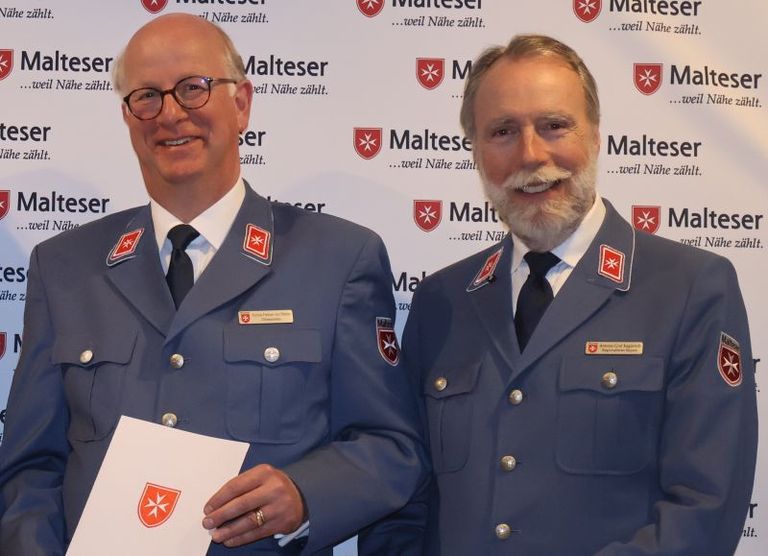 Zwei Männer in grauer Malteser Uniformjacke stehen vor einer weißen Wand mit vielen Malteser Logos