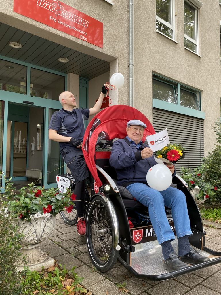 Ein Fahrgast sitzt in der Rikscha und der Ehrenamtliche Begleiter steht dahinter.