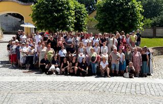 Ein Gruppenfoto von ca. 110 Erwachsenen. Im Hintergrund sind Bäume und eine Mauer zu sehen, die Sonne scheint.