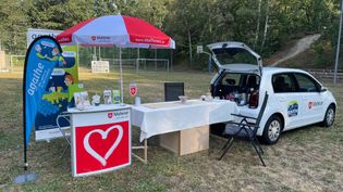 Weißer Malteser-Infostand mit rotem Herzlogo, rotem Sonnenschirm, blauem Agathe-Werbefahne und offenem weißen Auto auf Grasfläche.