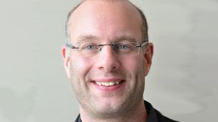 Nahaufnahme einer Person mit Brille vor hellem, neutralem Hintergrund.