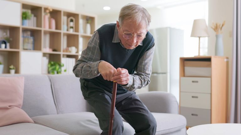 Ein alter Mann mit Gehstock hat Probleme, von einem Sofa aufzustehen.