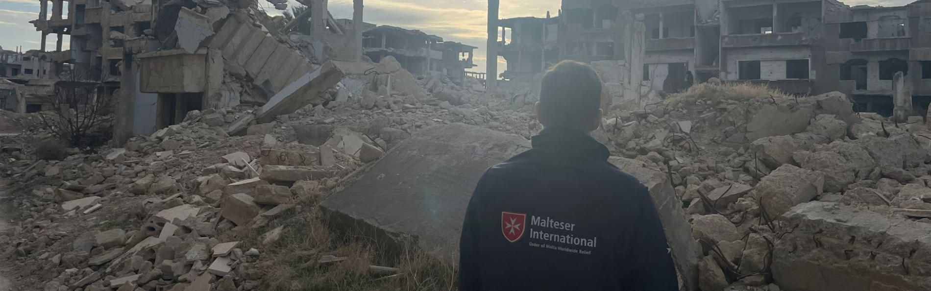 Ein Mann mit einer Jacke von „Malteser International“ steht mit dem Rücken zur Kamera vor einer Gebäuderuine in Syrien.
