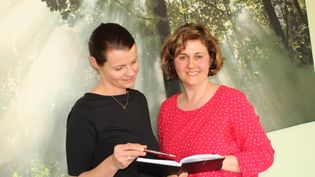 Zwei Frauen stehen vor einer Waldwand mit Sonnenstrahlen, eine trägt ein schwarzes Oberteil, die andere ein rotes Oberteil mit weißen Punkten, sie halten ein aufgeschlagenes Buch.