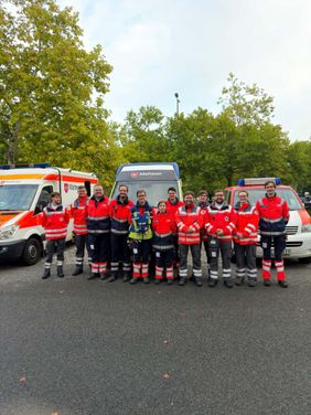 Auf dem Foto sind im Vordergrund mehrere Helfende der Übung seitens des DRK und der Malteser zu in Einsatzkleidung zu sehen. Im Hintergrund sind Einsatzfahrzeuge zu sehen.