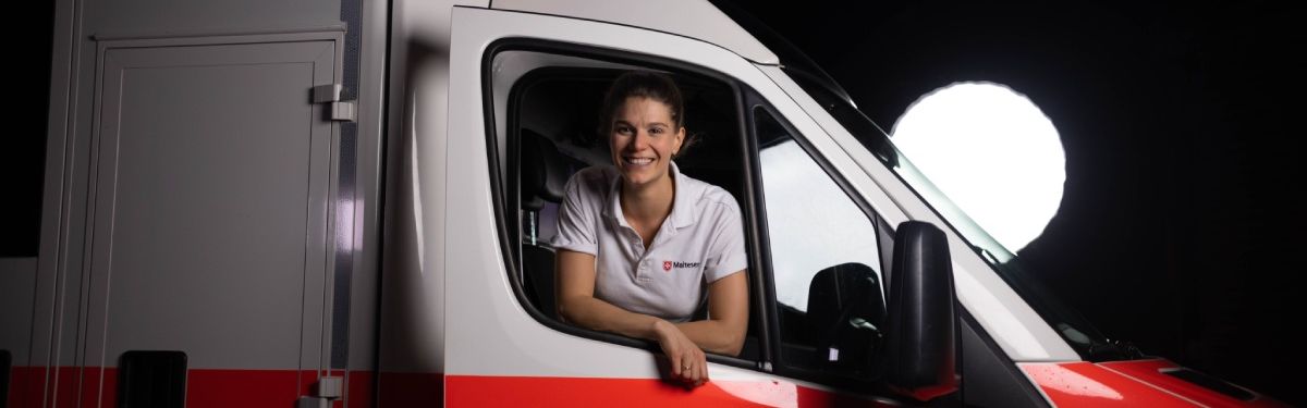 Eine junge Notfallsanitäterin schaut lächelnd aus dem Beifahrerfenster eines Rettungswagens.