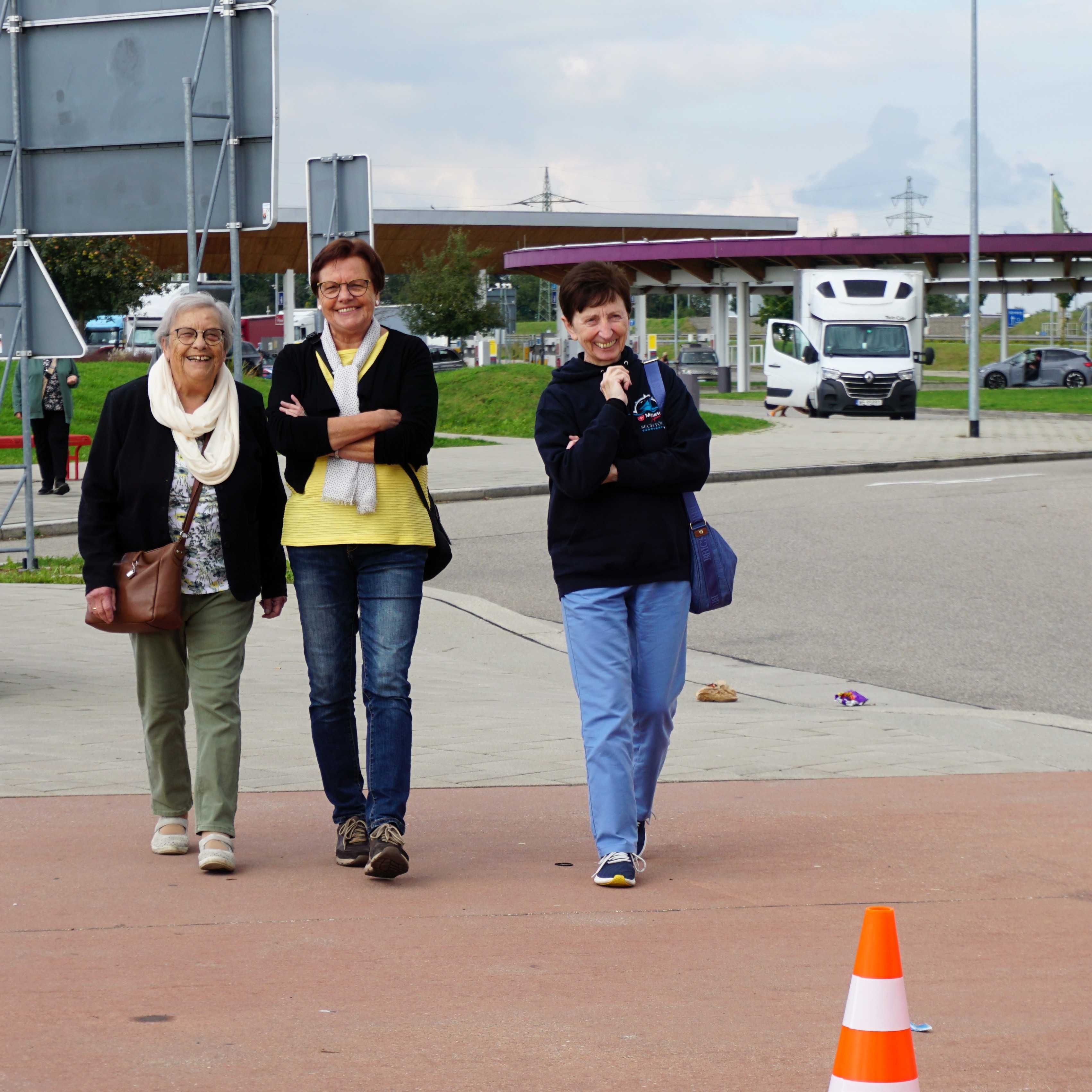 Drei Frauen gehen auf einem breiten Gehweg, im Hintergrund ein Lkw und ein überdachter Bereich.