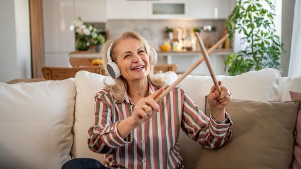 Eine ältere Frau mit Kopfhörern und Drumsticks sitzt lächelnd auf einem Sofa