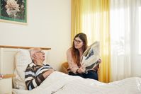 Eine junge Frau sitzt an einem Bett, in dem eine ältere Person liegt.