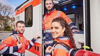 Zwei Rettungssanitäter und eine Rettungssanitäterin stehen in Schutzkleidung vor einem Rettungswagen.