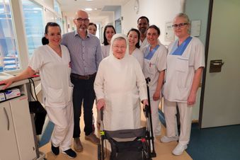 Ältere Frau mit Rollator im Klinikflur, umgeben von Pflegekräften und einem Arzt.