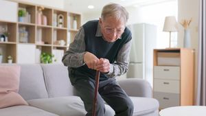 Ein alter Mann mit Gehstock hat Probleme, von einem Sofa aufzustehen.