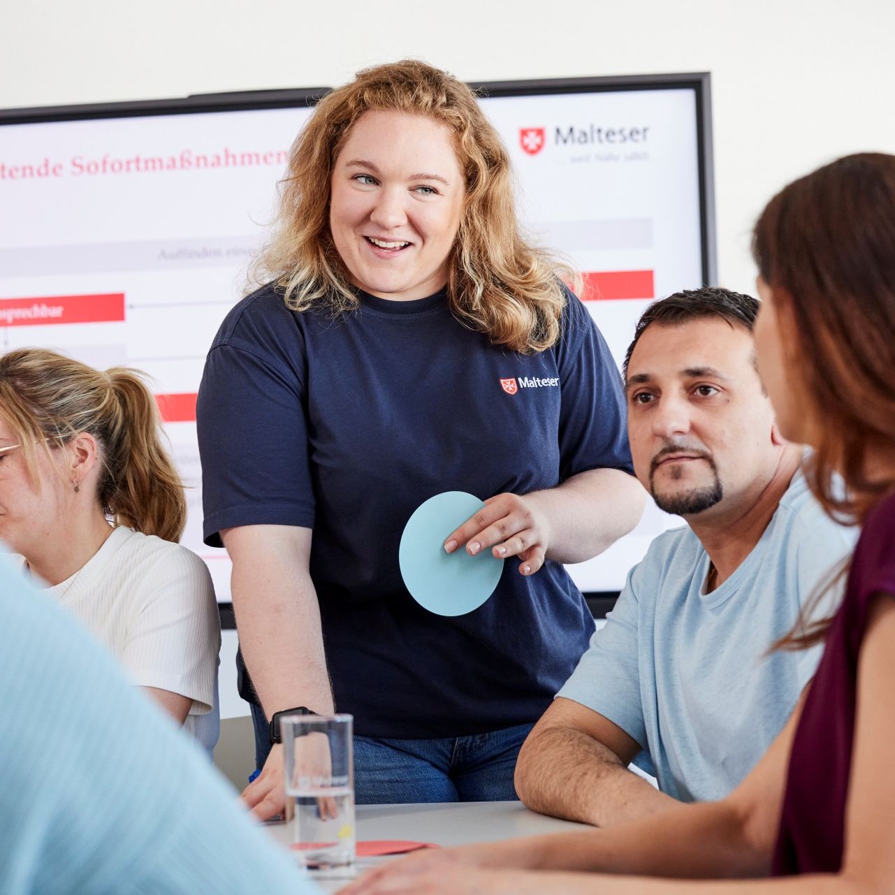Eine Person im Malteser Shirt erklärt in einer Schulungssituation Erste Hilfe, während mehrere Teilnehmende aufmerksam zuhören.
