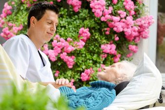 Ein junger Pfleger sitzt am Bett einer alten Frau. Das Bett steht auf der Terrasse.