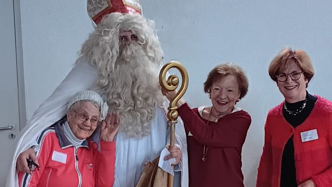 Eine große Person in traditionellem Nikolausgewand steht in der Mitte. Das Gewand ist weiß, dazu trägt die Person eine rote Mitra mit goldenen Verzierungen und hält einen goldfarbenen Bischofsstab in der rechten Hand. Über dem linken Arm hängt ein brauner Jutesack. Links und rechts vom Nikolaus stehen drei Senioren des Malteser Besuchsdienstes in festlicher Kleidung, die sich eng an ihn stellen. Die Gruppe befindet sich in einem Raum mit hellem Holzboden und weißer Wand im Hintergrund.