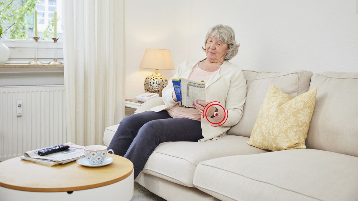 Kundin des Malteser Hausnotrufs trägt den Notfallknopf als Notrufarmband und ließt eine Zeitschrift auf dem Sofa.