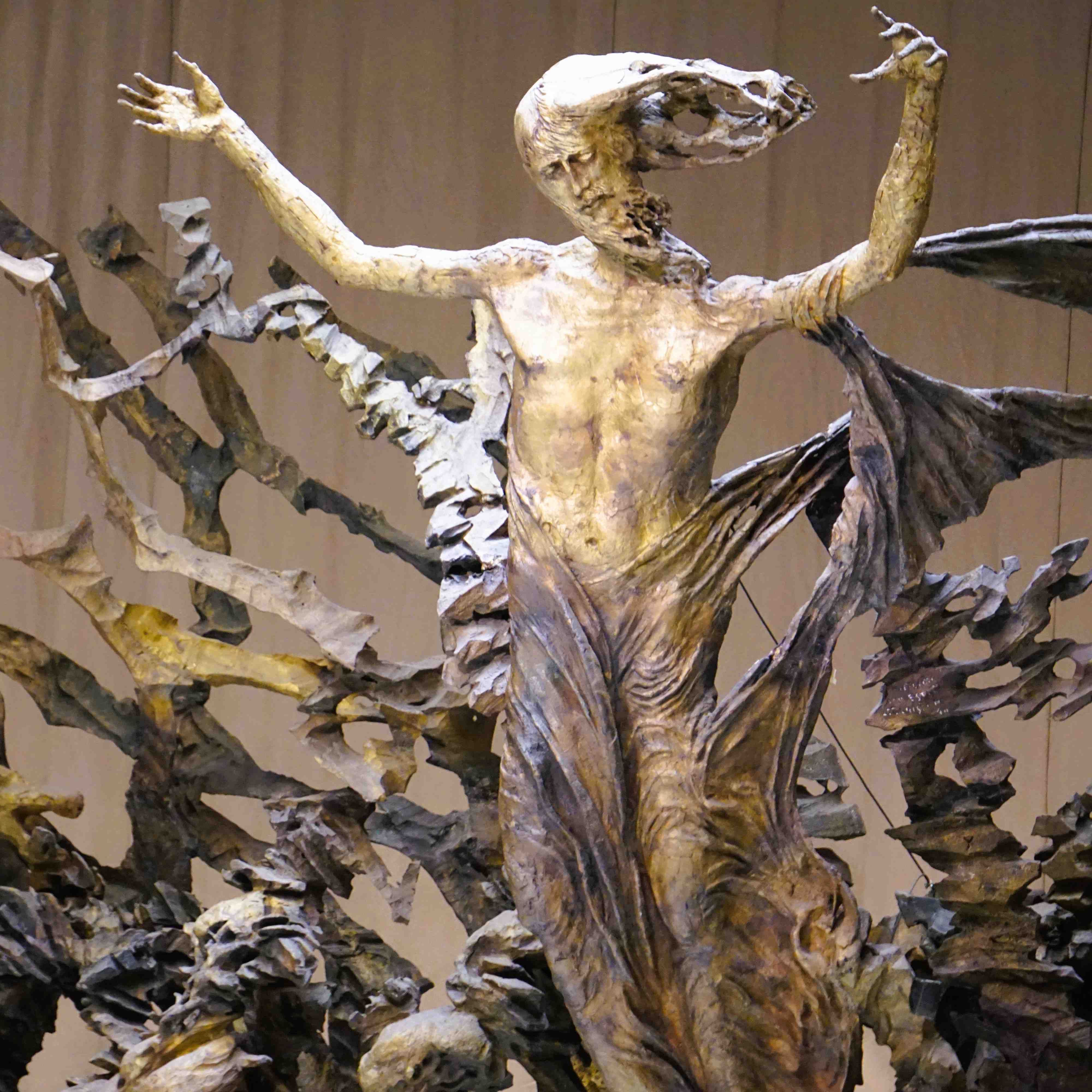 Skulptur eines aufrecht stehenden Mannes mit erhobenen Armen, umgeben von abstrakten, fliegenden Formen.