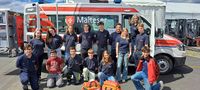 14 Schulsanis stehen oder knien gemeinsam vor einem Malteser Rettungswagen und schauen lächelnd in die Kamera. 