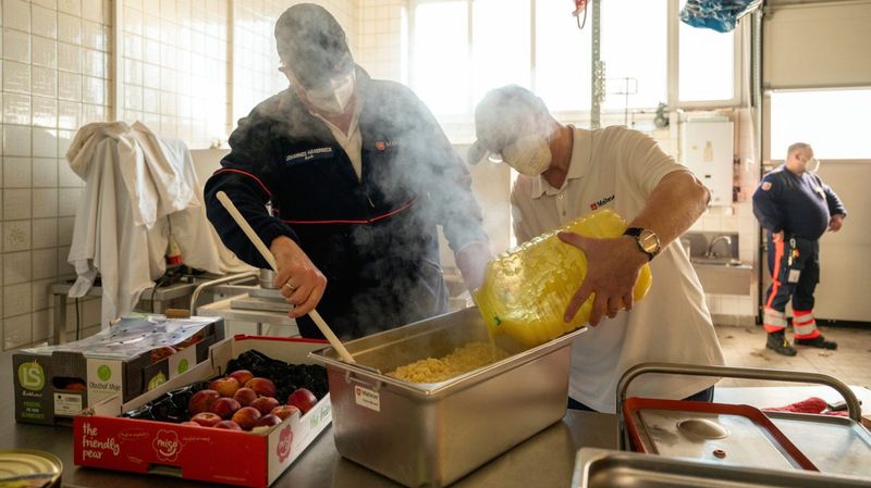 Zwei Männer mit FFP2-Masken bereiten Essen in einer Halle zu.