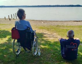 Eine Frau im Rollstuhl und ein Malteser Helfer, am Boden sitzend, schauen auf den See.
