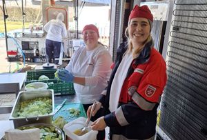 Zwei Frauen in Malteserkleidung bereiten Essen zu, vorne eine große Schüssel Gurkensalat, hinten vor dem Haus die Feldküche. 