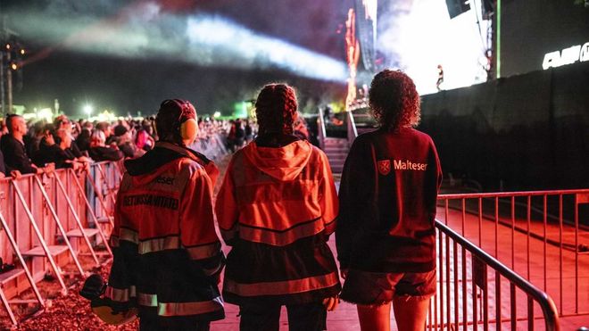 Rückansicht von drei jungen Frauen in Einsatzkleidung des Malteser Hilfsdienst im Bühnengraben eines Festivals. 