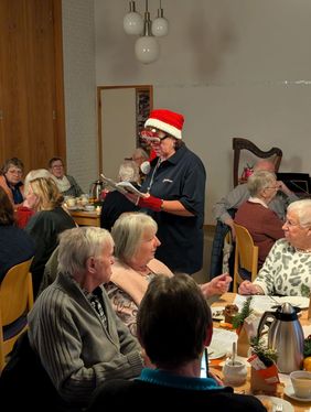 Eine Ehrenamtliche liest inmitten der Gäste der Weihnachtsfeier eine humrvolle Weihnachtsgeschichte vor und trägt dabei eine Nikolausmütze