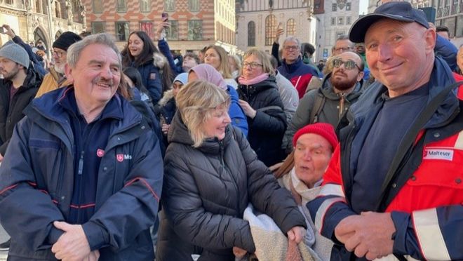 Ein Mann im nRollstuhl imitten von Maltesern und anderen Zuschauern auf dem Münchner Marienplatz.