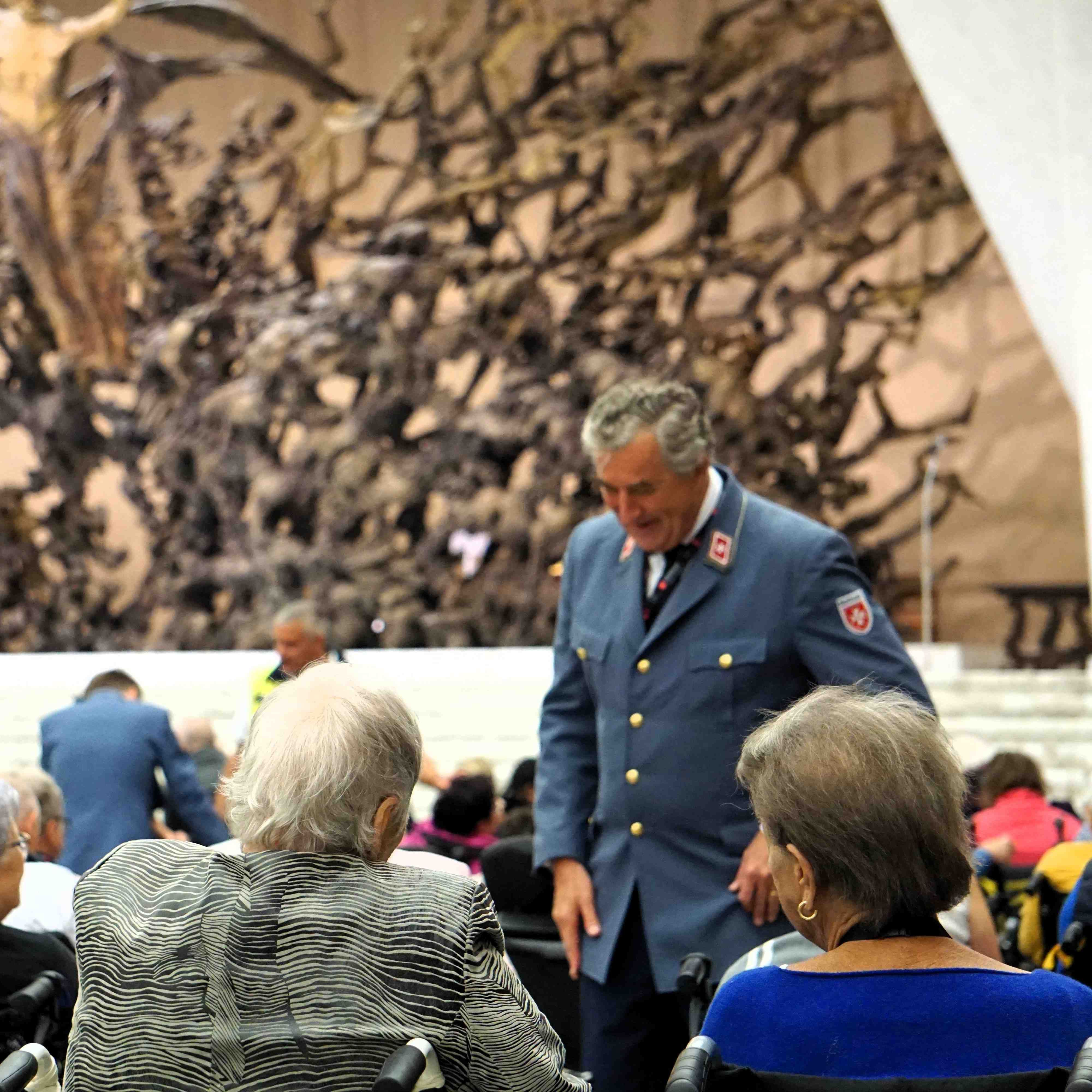 Mann in dunkelblauer Uniform steht vor Publikum, im Hintergrund große abstrakte Wandgestaltung aus Holz.
