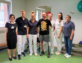 Patient mit Beinprothese steht in Physiotherapieraum, umgeben von sieben medizinischen Fachkräften und Angehörigen. 