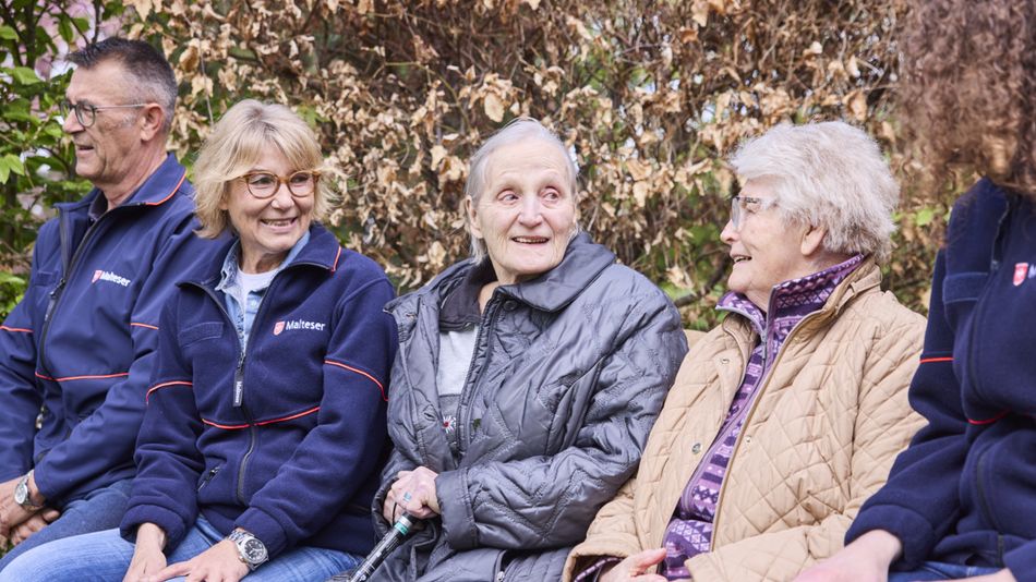 Mehrere ältere Menschen sitzen gemeinsam auf einer Bank im Freien. Zwei Personen in Malteser-Kleidung unterhalten sich mit anderen Teilnehmenden in einer herbstlichen Umgebung.