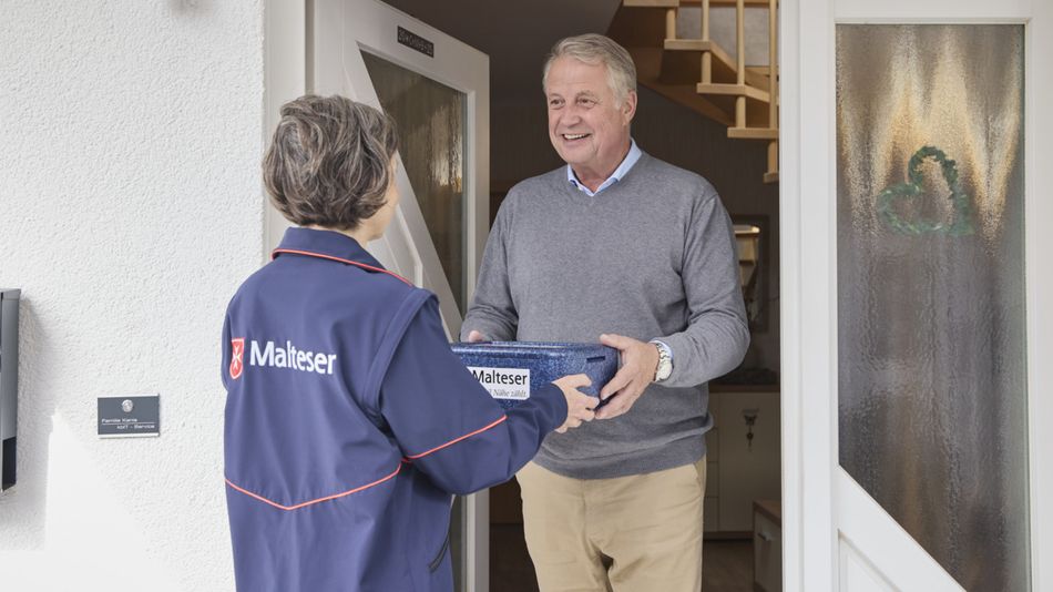 An einer Haustür wird gerade vom Malteser Menüservice das Essen übergeben