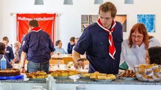 Leicht gebückte Frau in weißer Bluse und Mann in Malteser Kleidung stehen an einem Büffet. 