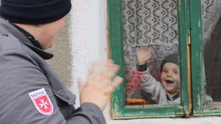Kleinkind mit Mütze winkt fröhlich durch ein Fenster mit Gardine, vor dem ein winkender Mann in Malteser Kleidung steht
