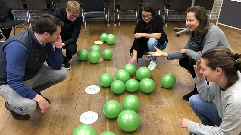 Mitarbeiter sitzen bei einem Workshop auf dem Boden vor grünen Kugeln. 