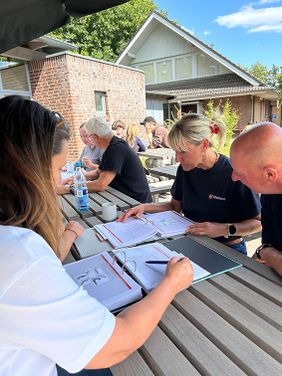 Mehrere Personen sitzen an Gartentischen auf der Terrasse und schauen in Lernmaterialien zur Ersten-Hilfe-Ausbildung.