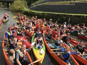 14 Kanus auf dem Wasser, in denen jeweils mehrere Personen sitzen und freudig in die Kamera lächeln.