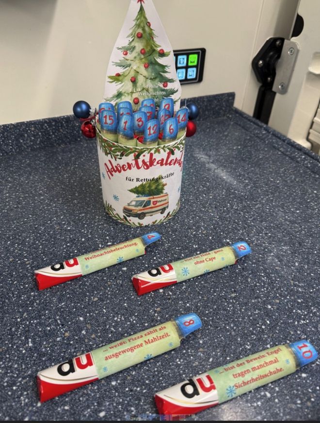 Ein selbst gebastelter Adventskalender für Rettungskräfte steht in einem Rettungswagen. Es handelt sich um eine Packung mit 24 Schokoriegeln. Auf jedem Schokoriegel befindet sind eine Banderole mit einem nummerierten Blaulicht und einer Du-Botschaft. 