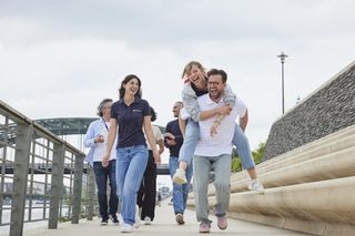 Eine Gruppe junger Menschen, jeglichen Aussehens und Geschlechts, spaziert lachend auf einer ausgebauten Flusspromenade entlang. Vorne wird eine Frau von einem Mann Huckepack getragen.