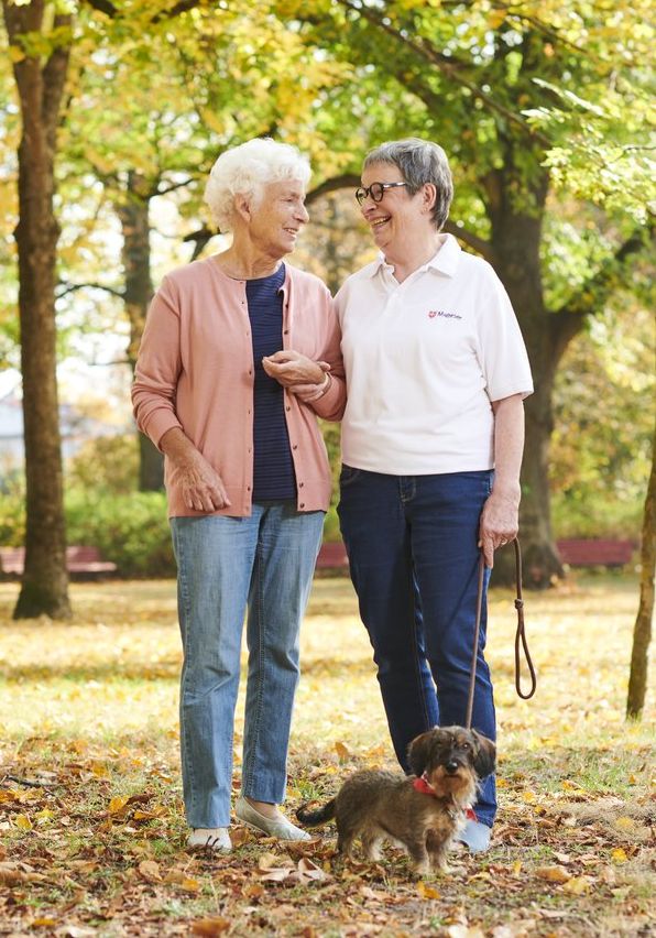 Eine ältere Dame wird von einer Ehrenamtlichen des Malteser Hilfsdienstes bei einem Spaziergang mit einem Dackel im Wald begleitet. Beide Frauen unterhalten sich und genießen die gemeinsame Zeit.