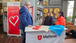 Ein Mann unterhält sich mit zwei Frauen. Sie stehen an einer Theke, auf der das Wort "Malteser" steht.