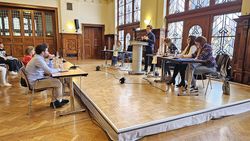 Menschen sitzen auf einer Bühne. Ein junger Mann steht an einem Pult und redet, andere hören zu.